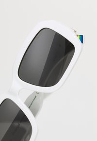 Lunettes de soleil blanches surdimensionnées avec des verres sombres, de forme carrée, et un accent multicolore distinctif sur la branche. Conception de cadre texturée.