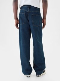 Jeans de mezclilla de pierna ancha en azul oscuro, con bolsillos traseros de parche y costuras en contraste, combinados con zapatillas negras con detalles en blanco.