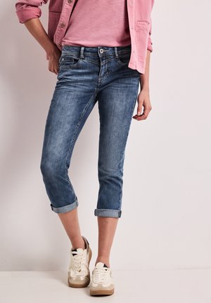 Femme portant un jean en denim bleu retroussé, un t-shirt rose pâle, une veste rose et des baskets blanches, debout devant un fond clair uni.