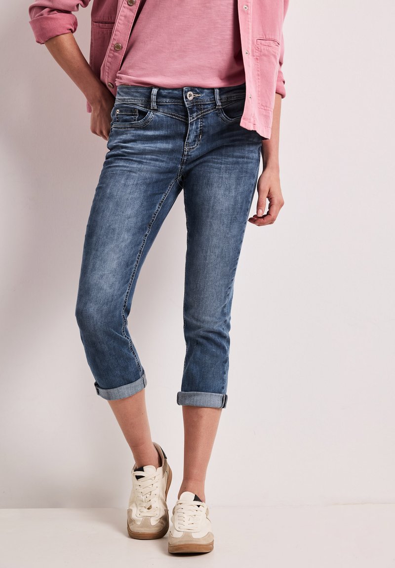 Donna con jeans blu risvoltati, t-shirt rosa pallido, giacca rosa e sneakers bianche, in piedi davanti a uno sfondo chiaro e uniforme.
