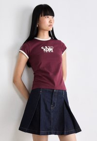 Burgundikangas t-paita valkoisella kauluksella ja hihoilla, jossa on Calvin Kleinin logo. Yhdistetty tummansinisestä farkkukankaasta valmistettuun hameeseen, jossa on laskoksia.
