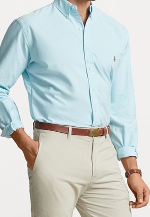 Uomo che indossa una camicia azzurra con bottoni, pantaloni beige, cintura di pelle marrone con fibbia dorata e cinturino dell'orologio in pelle marrone, mano in tasca.