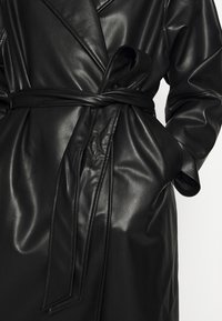 Trench-coat en cuir noir avec une taille ceinturée, de grands revers et une texture lisse et brillante, doté de poches latérales.