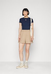 Marinblå kortärmad t-shirt med liten broderad design, matchad med beige veckade shorts. Modellen bär beige sneakers med krämfärgade strumpor och håller en tote-väska.