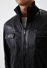 Czarna skórzana kurtka z wysokim kołnierzem, dwiema przednimi kieszeniami, pełnym zamkiem błyskawicznym i teksturowanym wykończeniem. Posiada kontrastowe szwy i żebrowany dekolt.