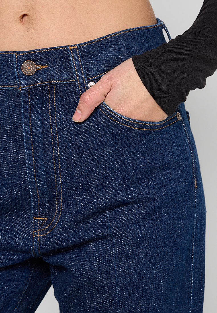 Jean en denim bleu foncé avec des coutures orange contrastantes, taille haute et une poche avant. Texture lisse et design classique avec une fermeture à bouton.