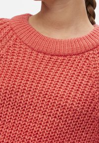 Zbliżenie na osobę noszącą teksturowany koralowy różowy sweter dzianinowy z ribbami w okrągłym dekolcie oraz widocznymi zaplecionymi włosami.