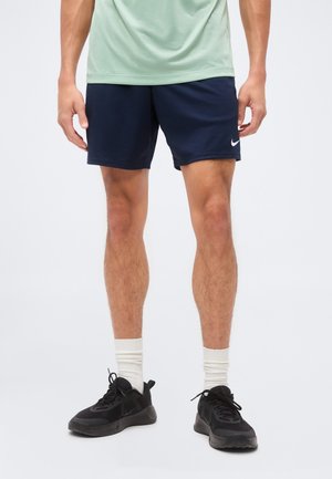 Hombre con pantalones cortos deportivos Nike azul marino, zapatillas negras, calcetines blancos y una camiseta verde claro, de pie frente a un fondo blanco.