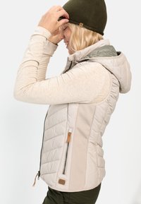 Beige Puffer-Weste mit Kapuze, ausgestattet mit gestepptem Textil, einer seitlichen Reißverschlusstasche und kontrastierender hellgrüner Innenauskleidung. Getragen über einem hellen Oberteil.