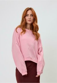 Világos rózsaszín, cropped pulóver, oversize ujjakkal, bordázott nyakkivágással és finom textúrával rendelkezik. Barna szoknya felett viselve.