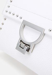 Hvid tekstureret læderhåndtaske med sølvfarvet hardware og nitter langs siden. Har en karakteristisk metallisk lukning med mærkegravering.