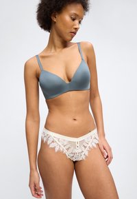 Γυναίκα φορά γκρι σουτιέν με λεπτές τιράντες και λευκά εσώρουχα από δαντέλα, ποζάροντας με το κεφάλι στραμμένο στο πλάι μπροστά σε μια απλή φόντο.