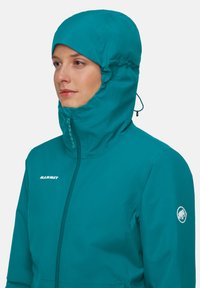 Giacca impermeabile di colore verde acqua con cappuccio, con zip frontale e logo sulla manica, progettata per attività all'aperto.