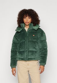 Groene corduroy pufferjack met een hoge kraag, drukknoopsluiting en horizontale stiksels. Een klein logo is zichtbaar op de linkerborst.
