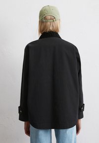 Czarna kurtka z klasycznym kołnierzem, z długimi rękawami i zapięciem na guziki przy mankietach. Widok z tyłu ukazuje luźny krój i minimalistyczny design.