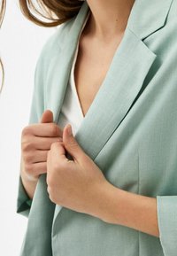 Žena upravující světle zelený blazer přes bílý texturovaný top, zobrazení detailu rukou a horní části trupu na bílém pozadí.