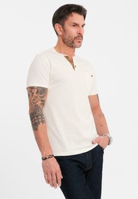 Kremowa koszulka z krótkim rękawem i zapięciem na guziki, z kieszonką na lewej piersi i brązowymi akcentami. W zestawieniu z ciemnymi dżinsami, z widocznymi tatuażami na ramieniu.