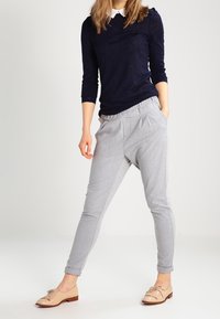 Camicia a maniche lunghe blu navy abbinata a pantaloni grigi chiari con vestibilità ampia e fascia elastica in vita. Scarpe beige con dettagli decorativi.