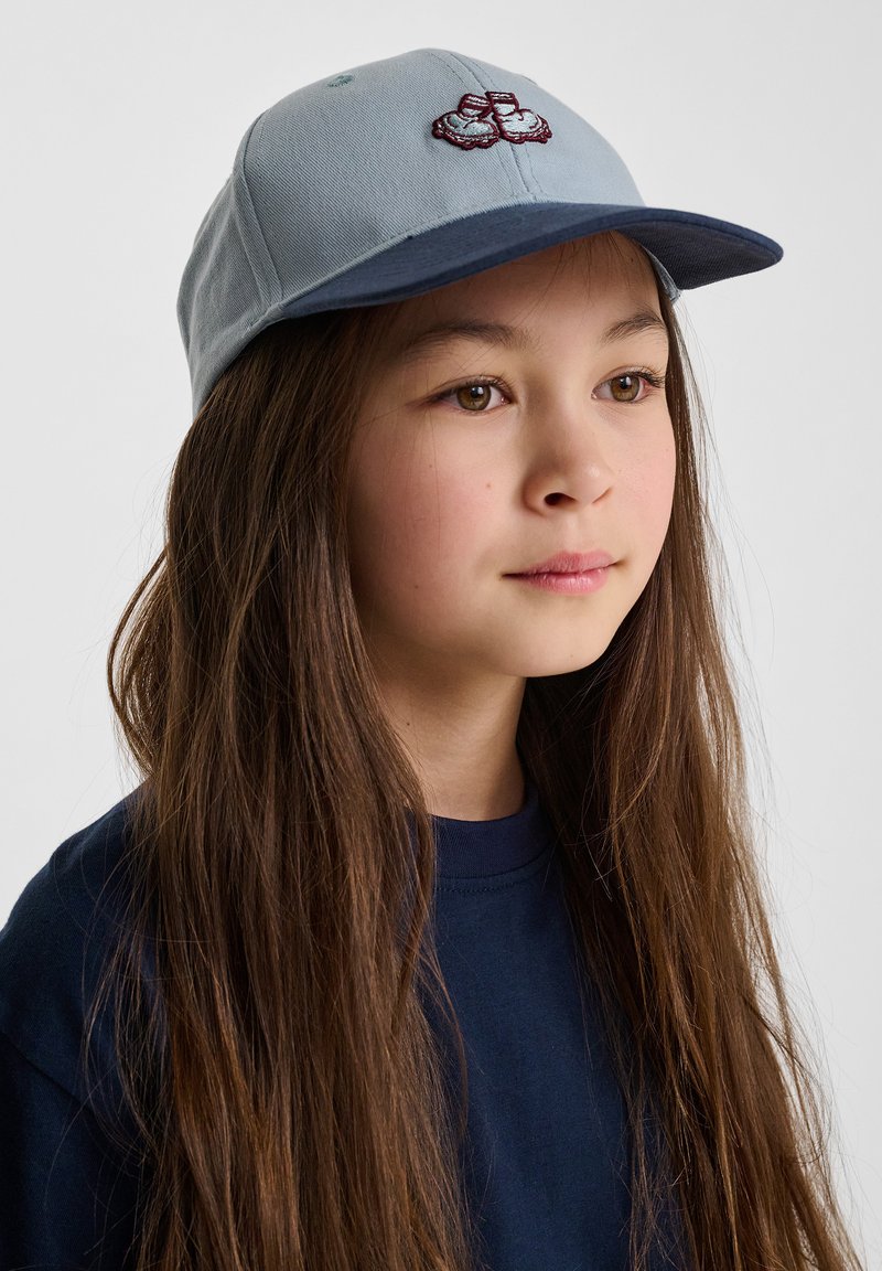 Ljusblå mössa med marinblå skärm, med en broderad design på framsidan. Tillverkad av bomull, strukturerad form och justerbar bakdel.