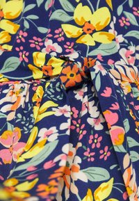 Květinové šaty s námořnickým pozadím, které zdobí jasně žluté, růžové a bílé květiny. Materiál je lehký a šaty mají detail vázání v pase.