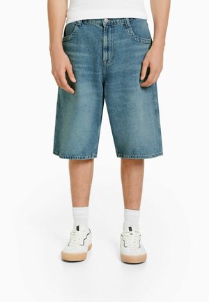 Hellblaue Jeansshorts mit lockerem Schnitt, knielang, ausgestattet mit vorderen Taschen und einem klassischen Knopfverschluss. Kombiniert mit weißen Sneakers.