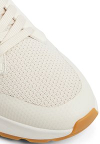Zapatilla con parte superior de malla texturizada en crema, punta de goma suave y suela de color beige claro. Presenta un cierre de cordones planos y un cuello acolchado.