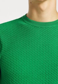 Grön texturerad stickad tröja med rund hals. Mönstret har en upphöjd diamantliknande design och ribbad krage.