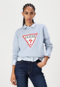 Felpa blu chiaro con un grande logo triangolare rosso con la scritta "GUESS", indossata sopra una camicia a righe blu e bianche con colletto e jeans scuri.