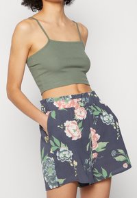 Žena v zeleném zkráceném topu a námornických květovaných šortkách se rukama v kapsách na jednotném pozadí.