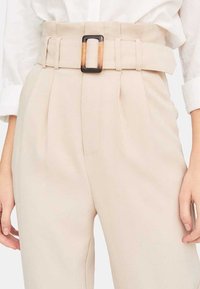 Béžové kalhoty vysokého pasu s předními sklady, vybavené širokým opaskem s obdélníkovou černohnědou sponou. Hladká textura tkaniny.