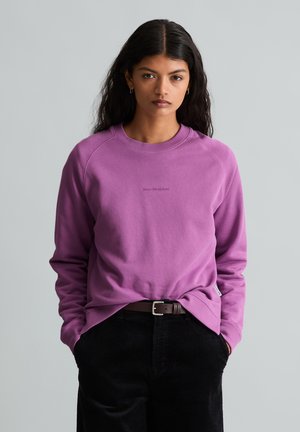Jonge vrouw met lang donker haar draagt een paarse sweatshirt en zwarte broek, staat met handen in de zakken tegen een effen achtergrond.