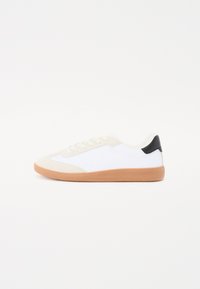 Fehér sneaker bézs velúr kiegészítőkkel, fekete saroktabbal, gumi talppal és perforált részletekkel az oldalán; alacsony profilú design.