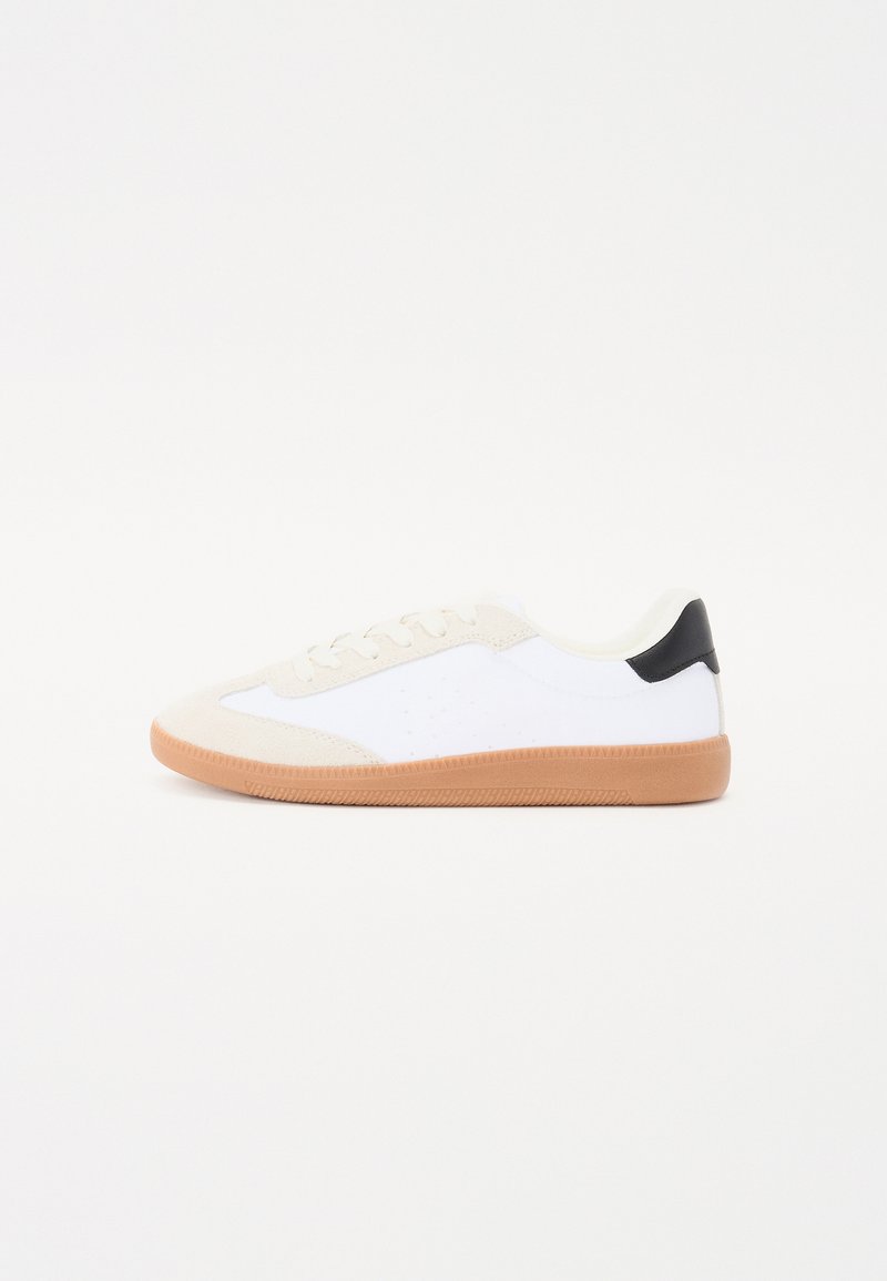 Fehér sneaker bézs velúr kiegészítőkkel, fekete saroktabbal, gumi talppal és perforált részletekkel az oldalán; alacsony profilú design.