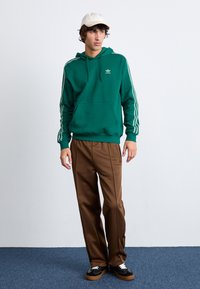 Grønn hettegenser med hvite striper og logo, kombinert med brune bukser og svarte joggesko. Modell med en kremfarget caps. Enkel bakgrunn.