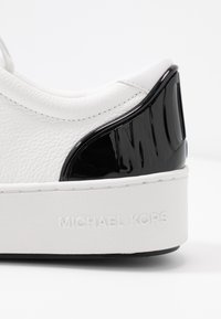 Baskets blanches en cuir texturé, accent en cuir verni noir, logo en relief, et semelle plate blanche avec une adhérence subtile.