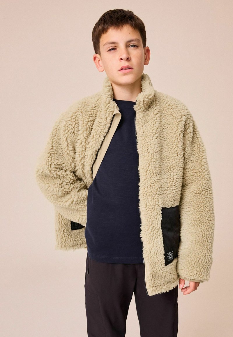 Beige Fleecejacke mit strukturierter Oberfläche, ausgestattet mit einer schwarzen Akzenttasche und einem Stehkragen, über einem marineblauen Shirt getragen.