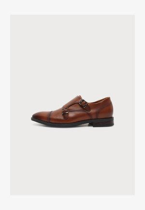 Brauner Monkstrap-Lederschuh mit zwei Metallschnallen, niedrigem Absatz und schwarzer Sohle, seitlich auf weißem Hintergrund gezeigt.