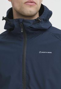 Mornarsko modra nepremočljiva jakna z visokim ovratnikom, prilagodljivo kapuco in črnim zadrgo. Na prsih ima logotip "North Bend".