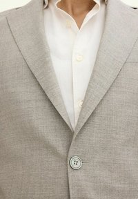 Blazer grigio chiaro con rever a punta, chiusura con un bottone e una leggera texture, abbinato a una camicia bianca con colletto e bottoni visibili.