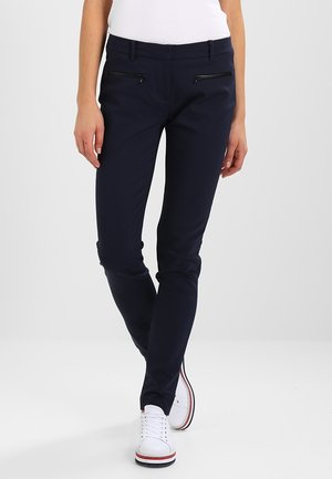 Persona que lleva pantalones ajustados de color azul marino oscuro con bolsillos negros con cremallera y zapatillas blancas con rayas rojas y negras en la suela.
