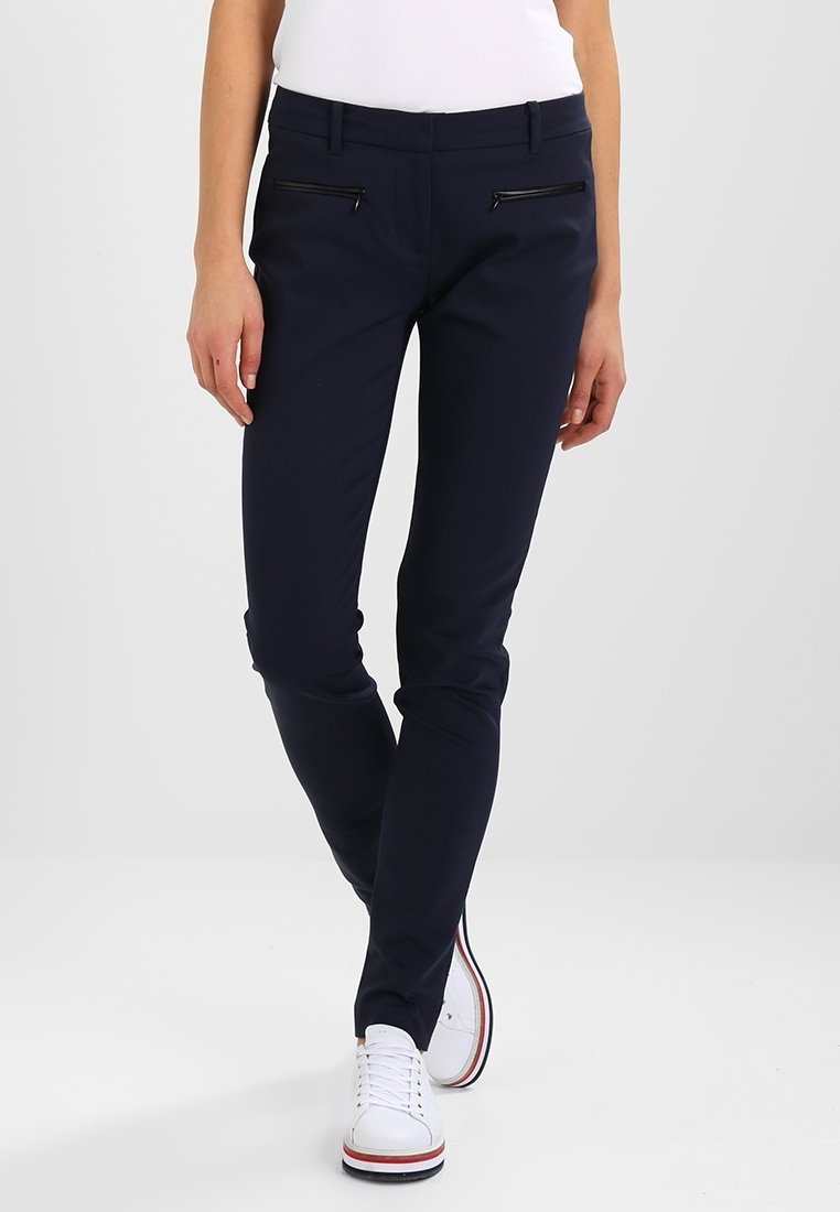 Personne portant un pantalon slim bleu marine foncé avec des poches zippées noires et des baskets blanches avec des bandes rouges et noires sur la semelle.