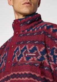 Polarowy pulower w burgundowym kolorze z granatowym geometrycznym wzorem. Posiada zapięcie na pół guzika, kieszeń zapinaną na zamek oraz logo Columbia z przodu.