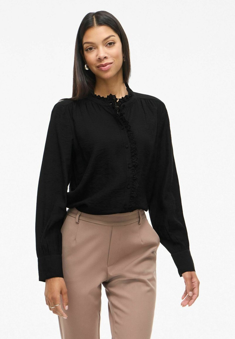 Melnā blūze ar garām piedurknēm, kurai ir riepšana priekšpuses plāksne un pogu aizdare. Valkāta ar gaiši brūnām piegriezuma biksēm. Gluda tekstūra.