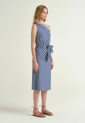 Donna che indossa un vestito midi monospalla con motivo blu e bianco, cintura in vita annodata e sandali bassi marroni, in piedi di profilo.