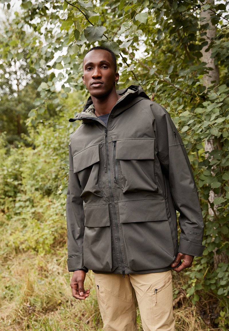 Chaqueta impermeable verde oliva con múltiples bolsillos frontales, cierre de cremallera y capucha ajustable, combinada con pantalones cargo beige en un entorno natural.