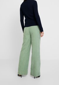 Person i svart räfflad långärmad topp och ljusgröna vida kordbyxor står mot en enfärgad ljusgrå bakgrund.