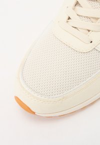 Light beige sneaker with a mesh upper, textured fabric, round toe, and orange rubber sole. Features flat laces and side stitching details.