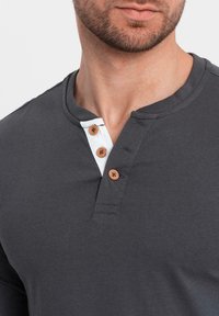 Mörkgrå långärmad tröja med rund halsringning, med en vit knapplist med tre bruna knappar för stängning och en slät bomullstextur.