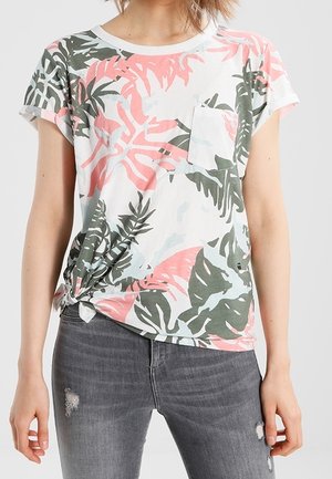 Femme portant un t-shirt blanc à manches courtes avec un imprimé de feuilles tropicales roses et vertes, noué à la taille, associé à un jean gris déchiré.