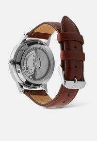 Rudas odinė apyrankė, nerūdijančio plieno korpusas, skaidrus korpuso dangtelis, atskleidžiantis mechanizmą, ir sidabro akcentai.